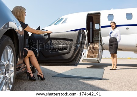 Elegant woman stepping out of car parked in front of private plane and airhostess