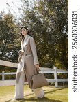 Elegant Woman in Stylish Beige Winter Coat Walking Outdoors with a Handbag, Surrounded by Bright Greenery and a White Fence.