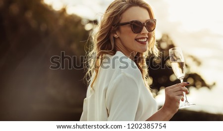 Elegant woman in sunglasses with a glass of wine outdoors. Smiling caucasian female having wine and looking backwards.