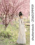 Elegant Woman in Traditional Chinese Hanfu Dress Posing Gracefully Amidst Blooming Pink Cherry Blossom Trees in a Serene Garden Setting.