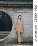 Elegant Woman in Traditional-Inspired Beige Outfit Posing by Ornate Circular Door in Tranquil Courtyard Setting.