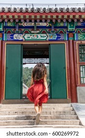 Elegant Woman Walking Through Old Wooden Traditional Chinese Gate At Ancient Imperial Temple, China Summer Travel. Gorgeous Girl With Flowing Hair And Fashion Red Dress In The Wind.