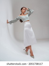 Elegant woman wearing pastel green top, silver silk skirt and high heels standing at white gray background and leaning on wall. Pretty fashion female model with pony tail brunette hair