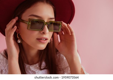 Elegant woman wearing trendy green rectangular sunglasses, dark red hat, pearl earrings, posing on pink background. Copy, empty space for text
