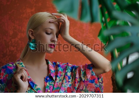 Elegant woman wearing trendy green shell earrings, colorful blouse, posing near orange wall. Summer fashion portrait. Copy, empty space for text