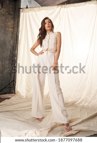 Elegant woman in white clothes standing against white textile background. Fashion portrait. Lady wearing palazzo pants and lace blouse