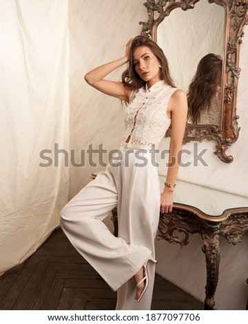 Elegant woman in white clothes standing leaning on marble table in bright interior against textile background. Fashion portrait. Lady wearing palazzo pants and lace blouse