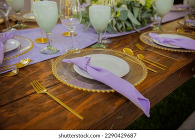 The elegant wooden table is decorated with gold linens and bright metallic accents in gold, green, and purple colors, topped with purple napkins and plates in a yellow and purple color scheme. The tab