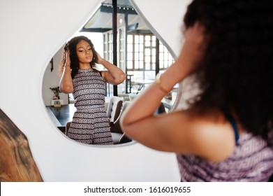 Сharming elegant young african american woman with long curly hair wearing jumpsuit looking at mirror.
