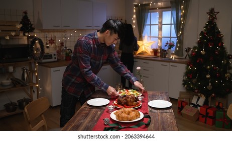 Elegant Young Asian Husband Arranging Silverware On Napkin On Dining Table At Home In Xmas Eve. Man Setting Up Table With Knife And Fork By Dishes While Girlfriend Preparing Delicious Cook In Kitchen