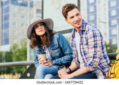 Elegant young brunette woman in hat and denim jacket and her boyfriend drinking coffee outdoor while waitig for bus or tram on stop with urban cityscape on background. Friendship, freedom concept. - Powered by Shutterstock