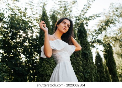 Elegant young brunette woman posing in white summer dress outdoors, low angle view. Beautiful slender girl with dark skin standing in fashion pose on background of trees. Beauty and fashion concept.
