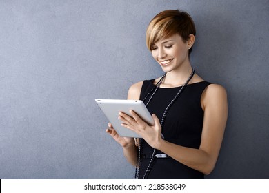 Elegant young businesswoman using tablet leaning to wall, looking at screen