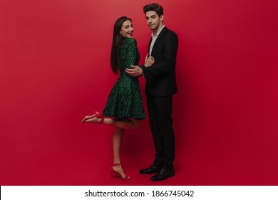 Elegant Young Couple In Green Dress And Black Suit Touching Each Other, Smiling And Posing Against Plain Red Background 