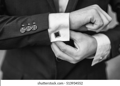 Elegant young fashion man looking at his cufflinks while fixing them. Black and white photo of male hands. Handsome groom dressed in black formal suit, white shirt and tie is getting ready for wedding - Powered by Shutterstock