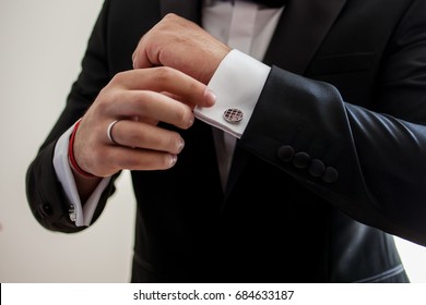 Elegant Young Fashion Man Looking At His Cufflinks While Fixing Them. Black And White Photo Of Male Hands. Handsome Groom Dressed In Black Formal Suit, White Shirt And Tie Is Getting Ready For Wedding