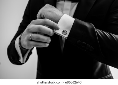 Elegant Young Fashion Man Looking At His Cufflinks While Fixing Them. Black And White Photo Of Male Hands. Handsome Groom Dressed In Black Formal Suit, White Shirt And Tie Is Getting Ready For Wedding