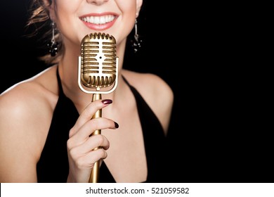 Elegant young female singer in black dress smiling holding golden vintage microphone, live performance, concert, unrecognizable person, close up, focus on mic, copy space. Black background