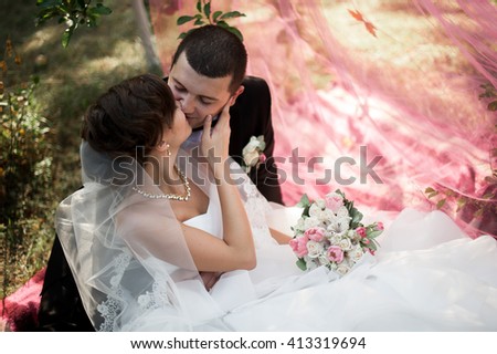 Elegant young happy wedding couple is sitting at green grass on green sunny summer background near the white canopy hugging, smiling and kissing.