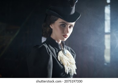 Elegant young lady in a 19th century suit poses in a dark vintage room full of smoke. Historical hairstyle and makeup.