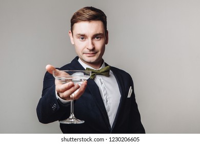 Elegant Young Man In Black Suit And Bow Tie Holding A Drink And Making Toast Gesture Isolated On Grey Background. New Year's Eve. Offering A Drink