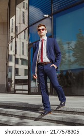 Elegant Young Man In Blue Suit, White Shirt, Red Tie, Blue Socks And Leather Shoes With Wooden Sunglasses At Contemporary Corporate Building