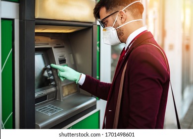Elegant young man with protective mask standing on city street and using ATM machin with protective gloves on hands. Virus pandemic prevention and healthcare concept.