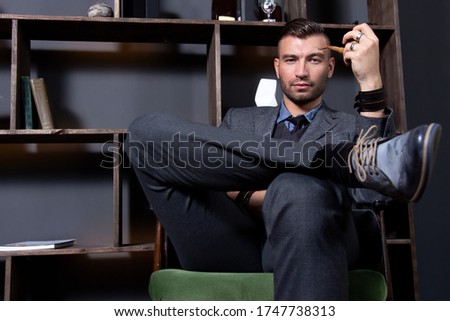 Elegant young man in a suit sitting, holding sigaret and serious watching. Relaxing in workoffice after hard day.Successful businessman