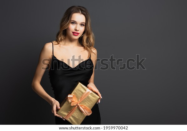 elegant young woman in black slip dress holding\
wrapped gift box isolated on\
grey