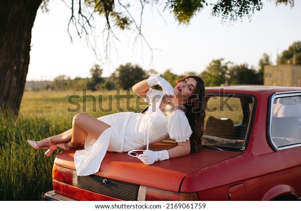 An elegant young woman with long curly hair\
in a white dress, white gloves and sunglasses lies on the trunk of\
a red retro car on a field\
road.