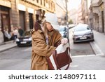 Elegant young woman looking in her brown and white bag her phone or purse. Traveler style woman wear brown trench coat, white cap, sweatshirt and bag on the street. Street style, fashion outfit.