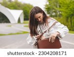 Elegant young woman looking in her brown bag her phone or purse. Traveler style woman wear jeans jacket, top and bag on the street. Street style, fashion outfit, woman walk in park, look happy.