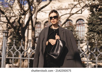 Elegant Young Woman Looking In Her Black Leather Bag Her Phone Or Purse. Business Style Woman Wear Grey Blazer, Black Eyeglasses And Bag On The Street. Street Style, Fashion Outfit.