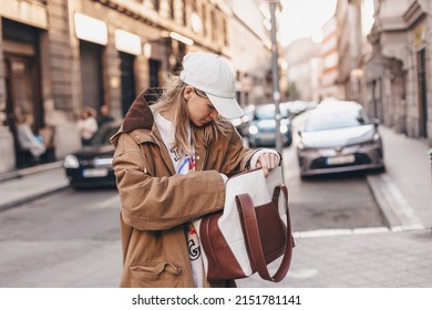 Elegant young woman looking in her brown and white bag her phone or purse. Traveler style woman wear brown trench coat, white cap, sweatshirt and bag on the street. Street style, fashion outfit.