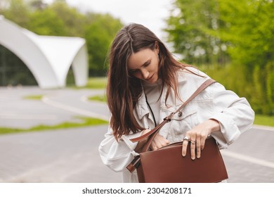 Elegant young woman looking in her brown bag her phone or purse. Traveler style woman wear jeans jacket, top and bag on the street. Street style, fashion outfit, woman walk in park, look happy.