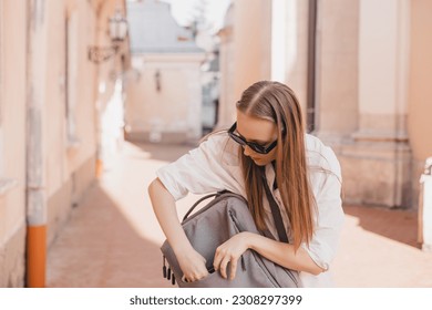 Elegant young woman looking in her grey backpack her phone. Traveler style woman wear white shirt, black top and glasses on the street. Girl walk on old city building, search her mobile to take photo.