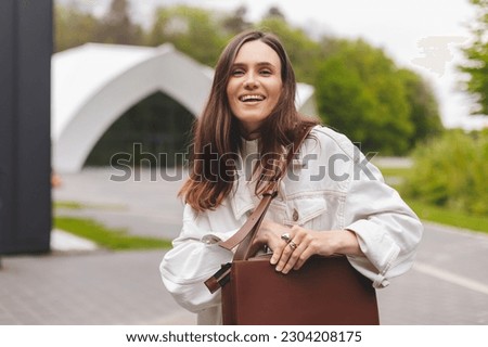 Elegant young woman looking in her brown bag her phone or purse. Traveler style woman wear jeans jacket, top and bag on the street. Street style, fashion outfit, woman walk in park, look happy.