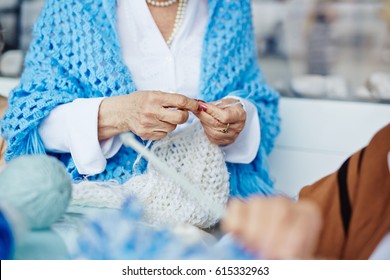 Elegant-looking senior woman in white shirt and cornflower blue shawl sitting indoors with unfinished knitted scarf on laps, colorful yarns lying on table