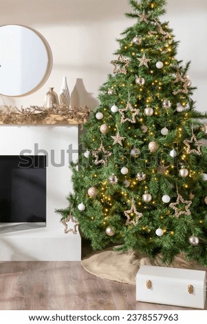 Elegantly decorated Christmas tree next to a modern fireplace, adorned with glittering gold stars, white baubles, lights, round mirror and contemporary vases set, wooden floor, in a Nordic-style room.