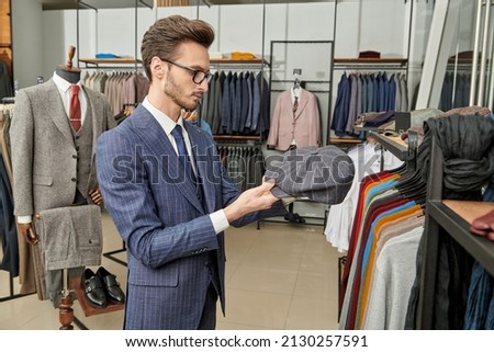 An elegantly dressed brunet man in spectacles chooses clothes in a premium men's clothing store. Men's business fashion. Sideview shot.