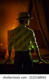 Elegantly Dressed Masculine Man  Stands With His Back To The Camera In A Dark Bedroom In Front Of The Table With Playing Cards And Banknotes. Detective Story, Mafia, Gambling.