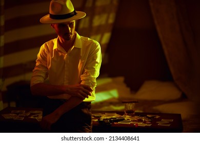 Elegantly Dressed Masculine Man  Stands Leaning On A Table With Banknotes And Playing Cards In A Dark Bedroom. Detective Story, Mafia, Gambling.