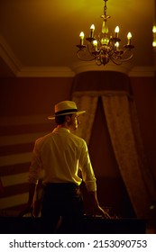 Elegantly Dressed Masculine Man  Stands With His Back To The Camera In A Dark Bedroom In Front Of The Table With Playing Cards And Banknotes. Detective Story, Mafia, Gambling.