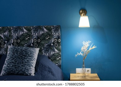 Element Of Cozy Bedroom In Modern Apartment With New Interior, Sconces Lamp On Blue Wall And Flowers At Nightstand Table Near Comfortable Bed