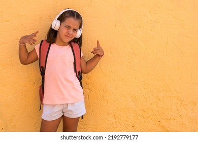 Elementary Age Smiling Girl Listening To Music With Wireless Headphones. Little Girl On Yellow Background. Positive Emotion. Back To School With Funny Gesture	