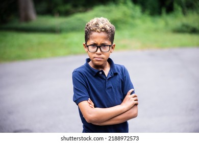 An Elementary Aged Boy With Glasses On And His Arms Crossed With A Mad Or Mean Look On His Face.