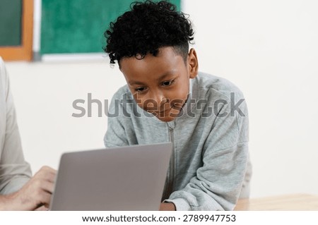 Elementary boy smiles while using laptop during teacher supported classroom task, building digital learning confidence, classwork routine, attention, problem solving, school readiness, and progress