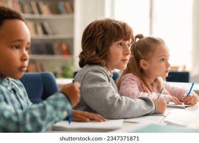 Elementary classroom of diverse children listening attentively to their teacher giving lesson, kids in school writing in exercise notebooks or taking a test