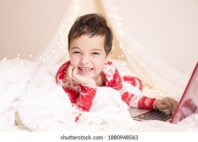 Elementary School Child On Winter Break During A Pandemic. Cheerful Kid In Red Christmas Pajamas With A Laptop In Bed. Concept Of Stay At Home, Family Holidays And Online Greetings. Horizontal. 