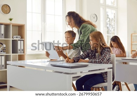 Elementary school children doing test in class. Group of kids sitting at desks in classroom and writing. Woman teacher helps student boy who doesn't understand something in his test